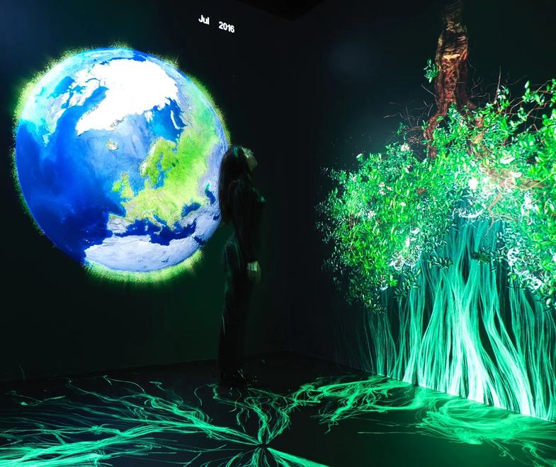 A person stands in Space for Earth, an immersive experience that is part of the Earth Information Center, surrounded by glowing digital projections of Earth and vibrant green foliage with flowing light patterns on the floor.