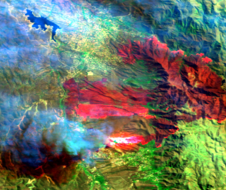 Satellite image showing an active wildfire, with bright red hot spots and smoke spreading over a mountainous landscape.