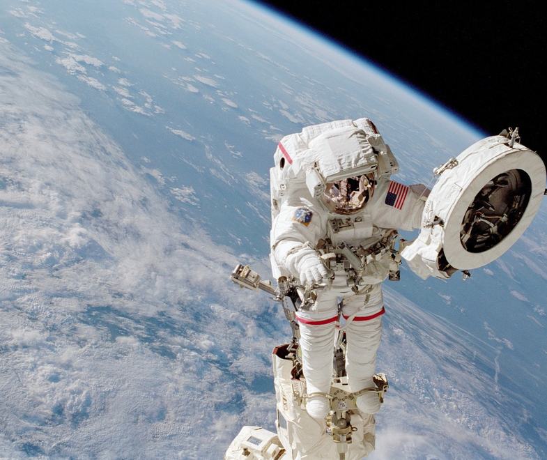 An astronaut performs a spacewalk above Earth, holding a large circular equipment panel while attached to the spacecraft.